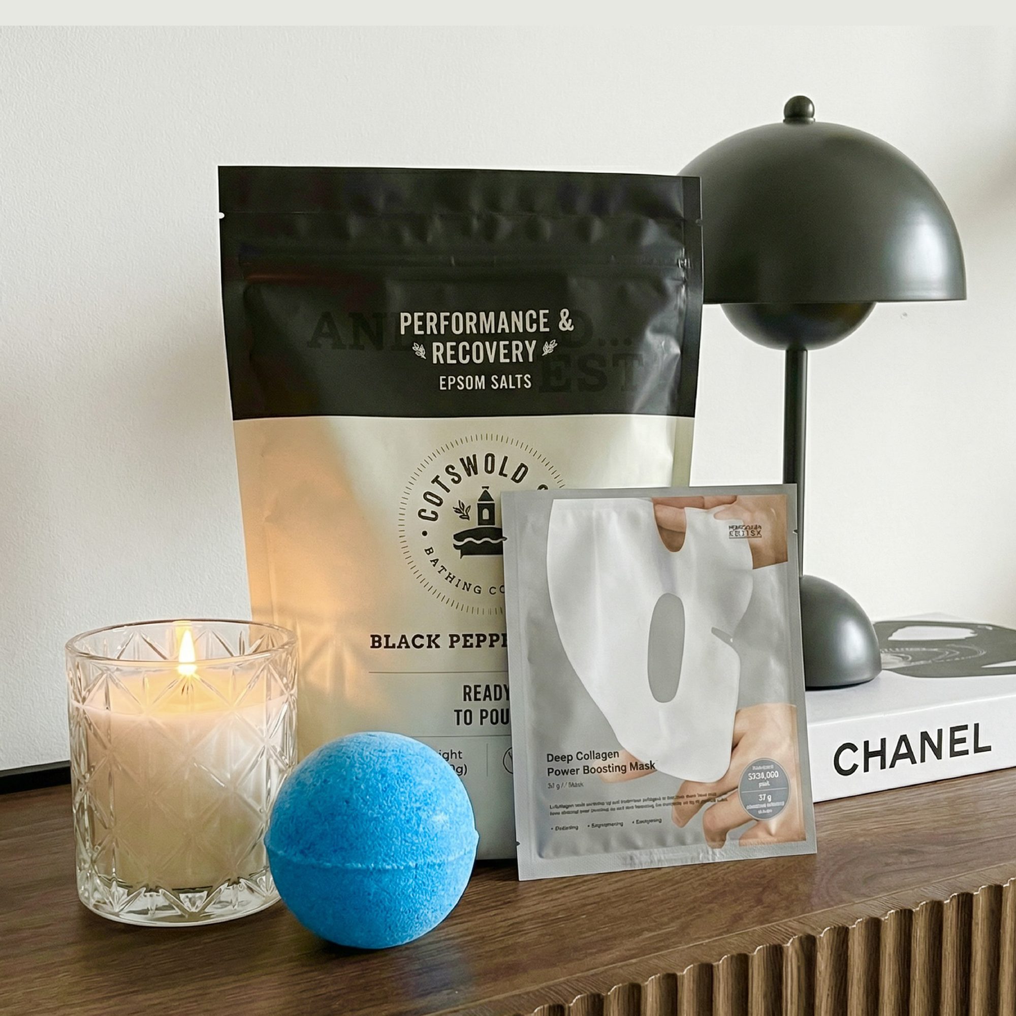 Spa and wellness products displayed on wooden surface including epsom salts, bath bomb, face mask, candle, and lamp