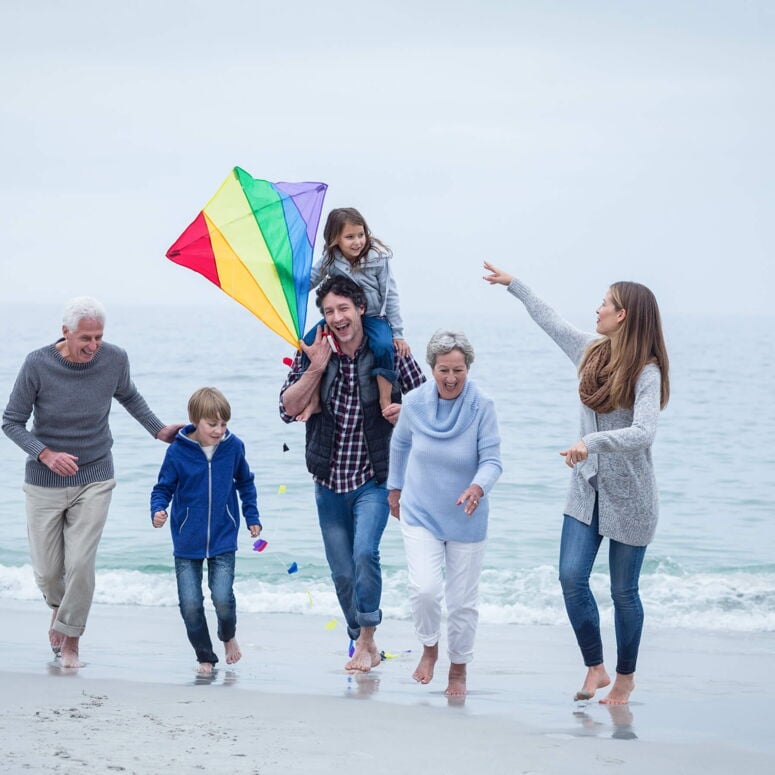 Familie mit Regenbogen-Drachen läuft barfuß durchs flache Wasser am Strand