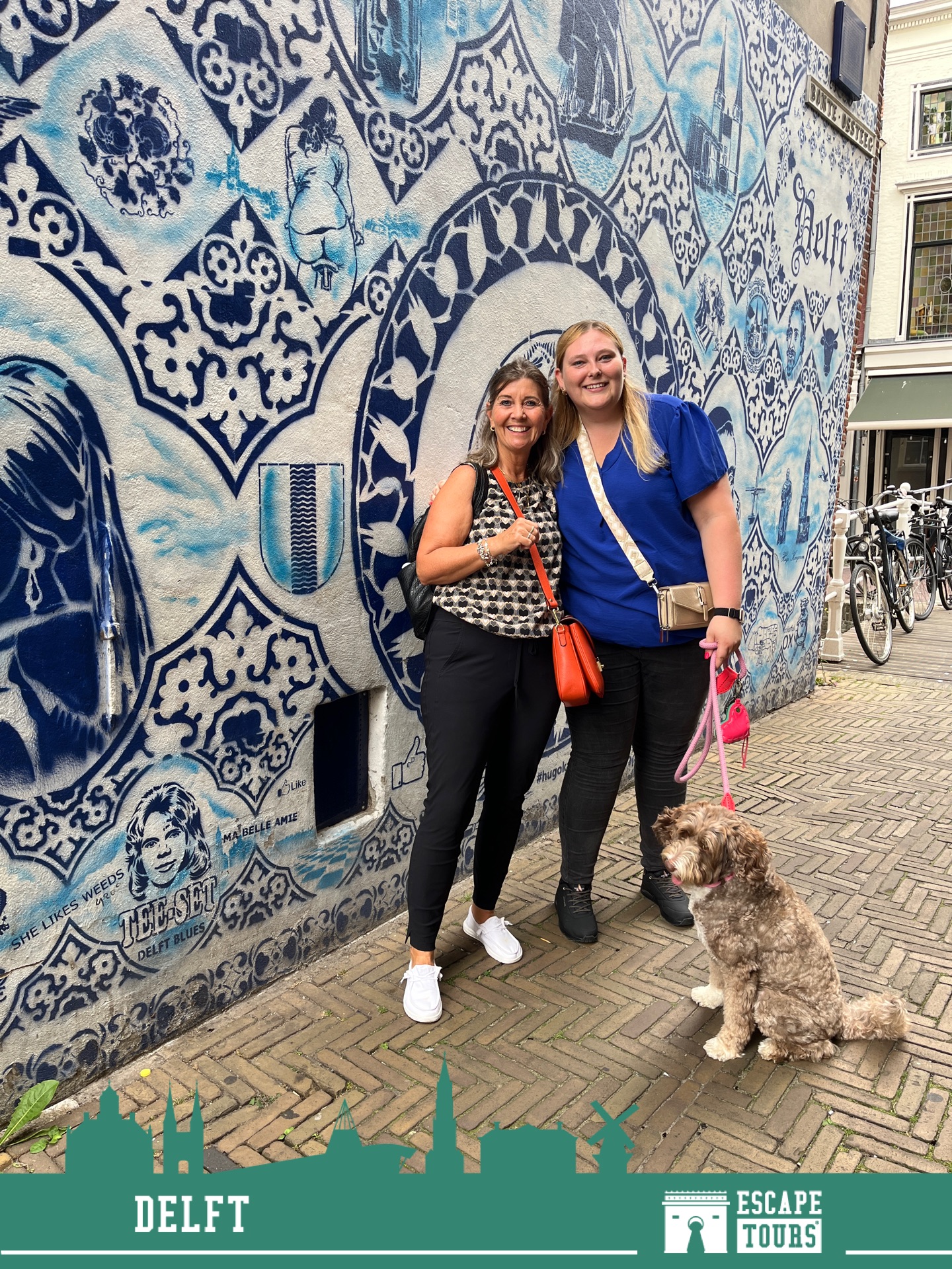 Two women posing in front of a large blue and white decorative mural with a small dog in the foreground