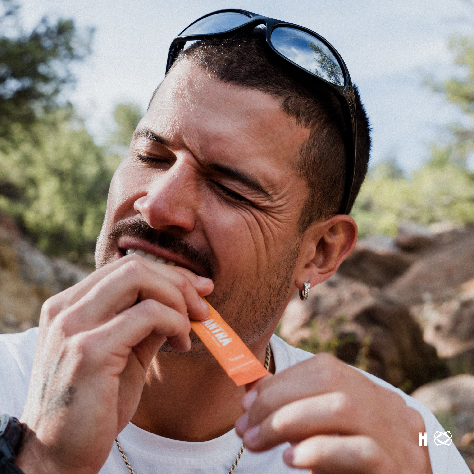 Man eating an orange-wrapped sachet outdoors with mountains and trees in background