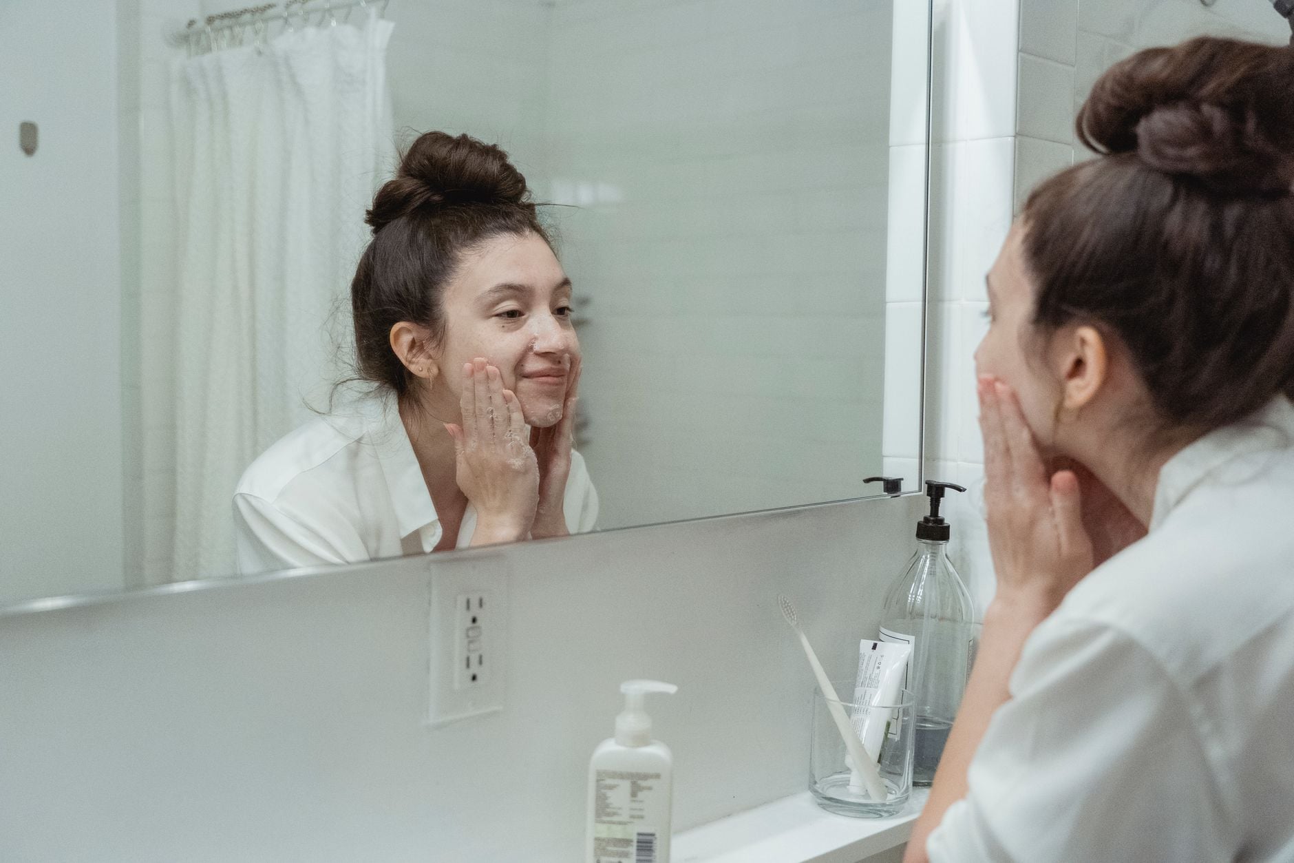 Woman examining her face in bathroom mirror while applying skincare product to her cheek