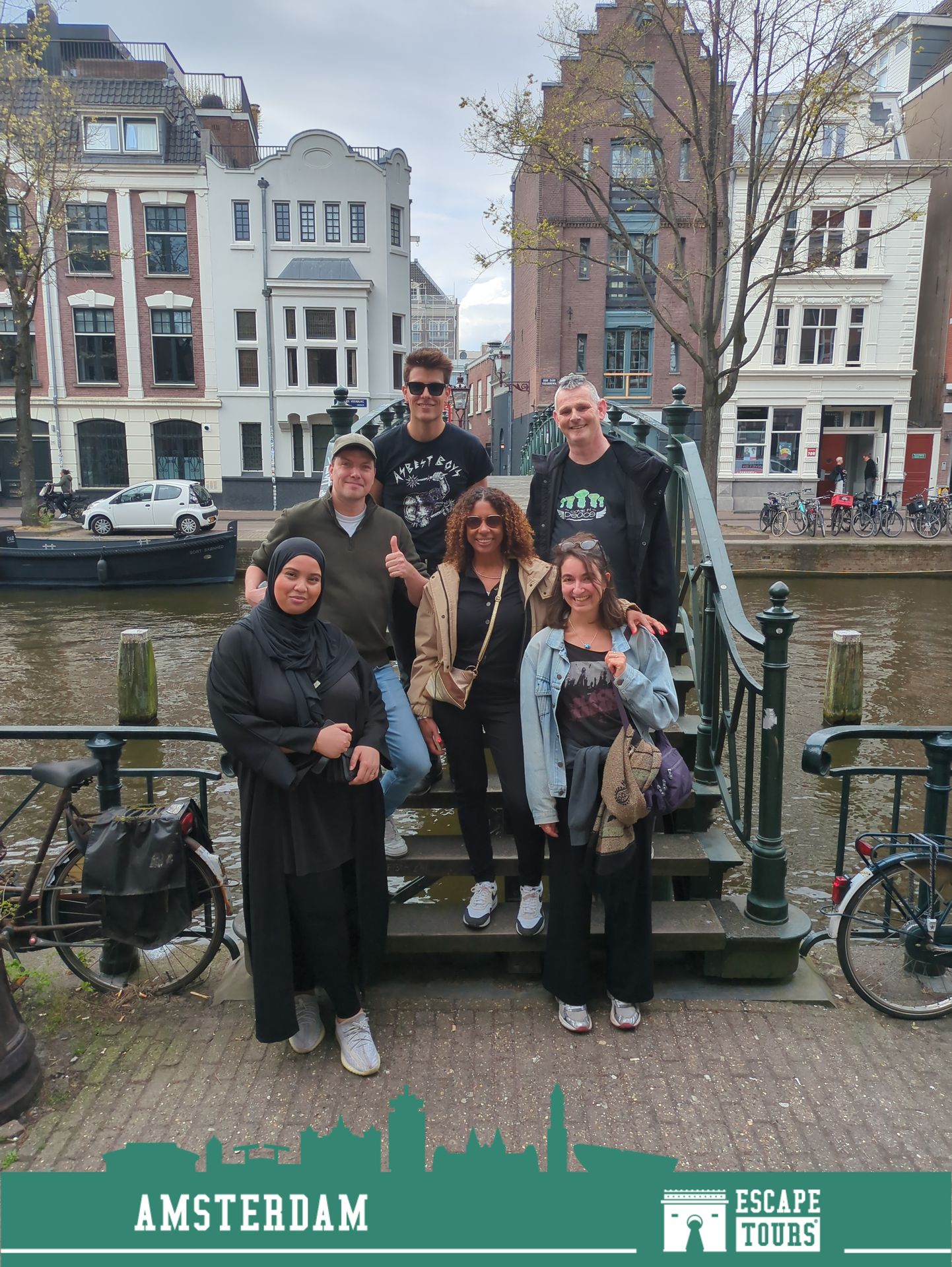 Group of six people posing together on a wooden dock by a canal with historic buildings in the background