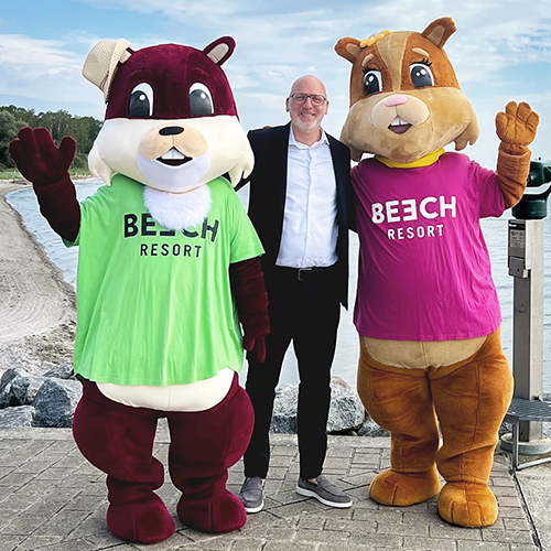 Mann in Anzug posiert mit zwei Maskottchen-Charakteren in bunten Shirts am Strand