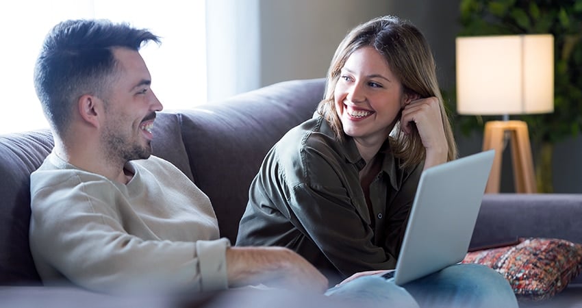 Man en vrouw zitten op grijze bank met laptop en kijken elkaar lachend aan