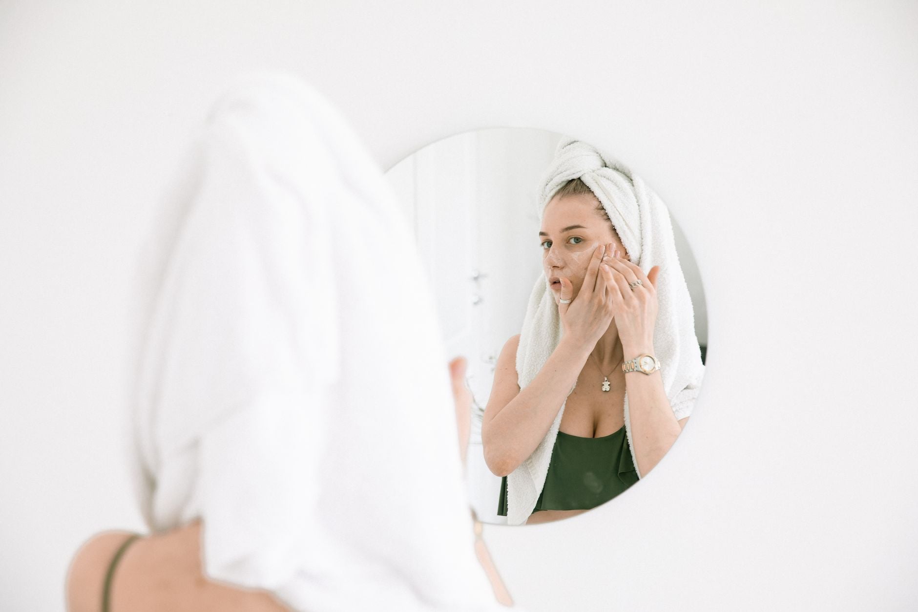 Woman in bathroom mirror with white towel wrapped around head, touching her face with concerned expression