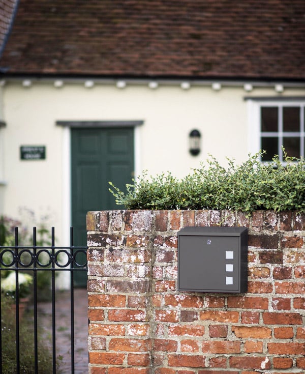 Dark gray security mailbox mounted on red brick wall with ivy, beside iron fence and cream-colored cottage