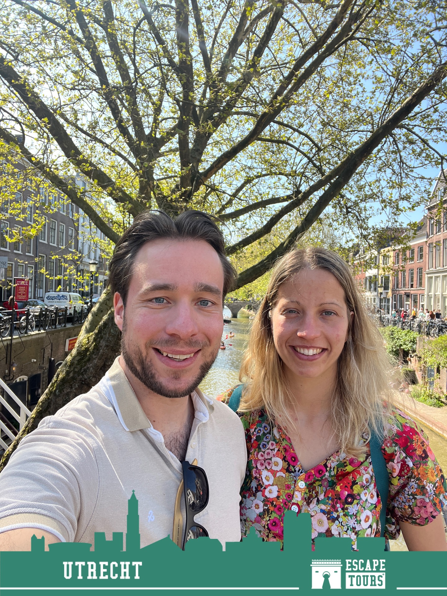 Couple posing together under a large tree by a canal with historic European buildings and crowds in the background